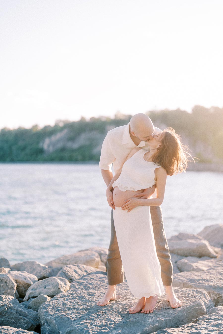 Expectant couple kissing on rocks beside Lake Ontario at Scarborough Bluffs.