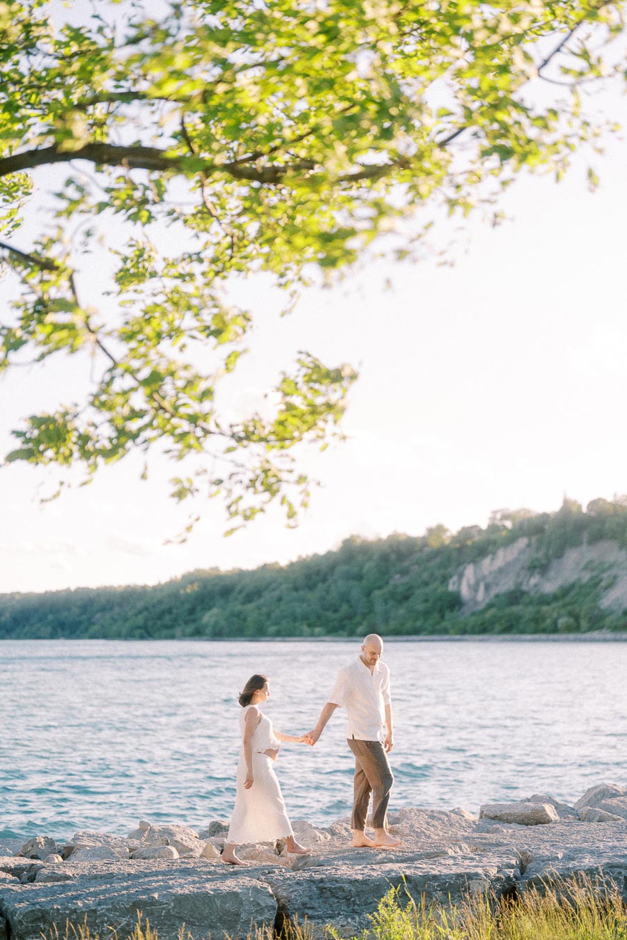 Pregnant couple holding hands while walking on rocks beside Lake Ontario at Scarborough Bluffs.