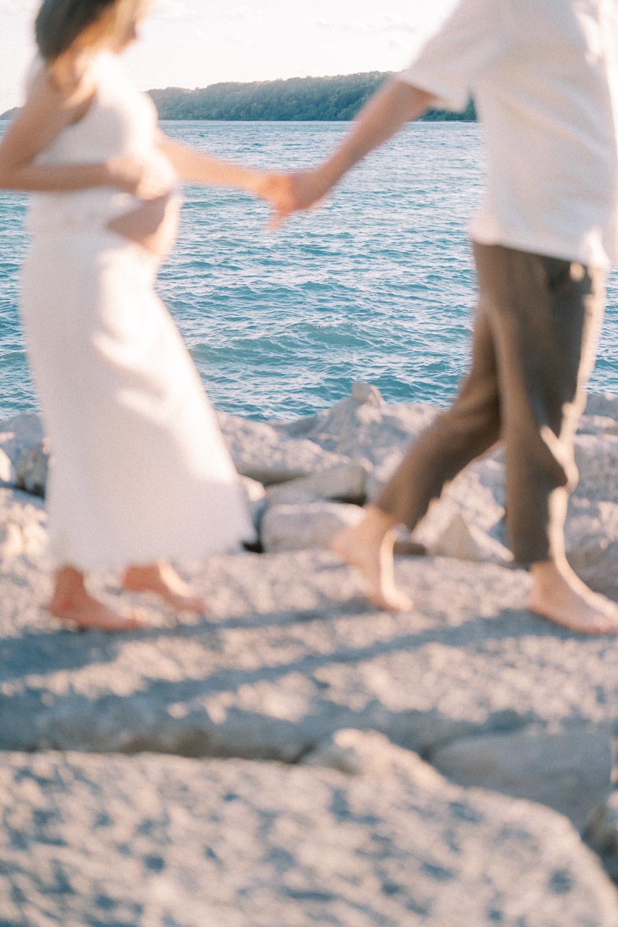 Expectant couple holding hands while walking barefoot on rocks beside Lake Ontario.