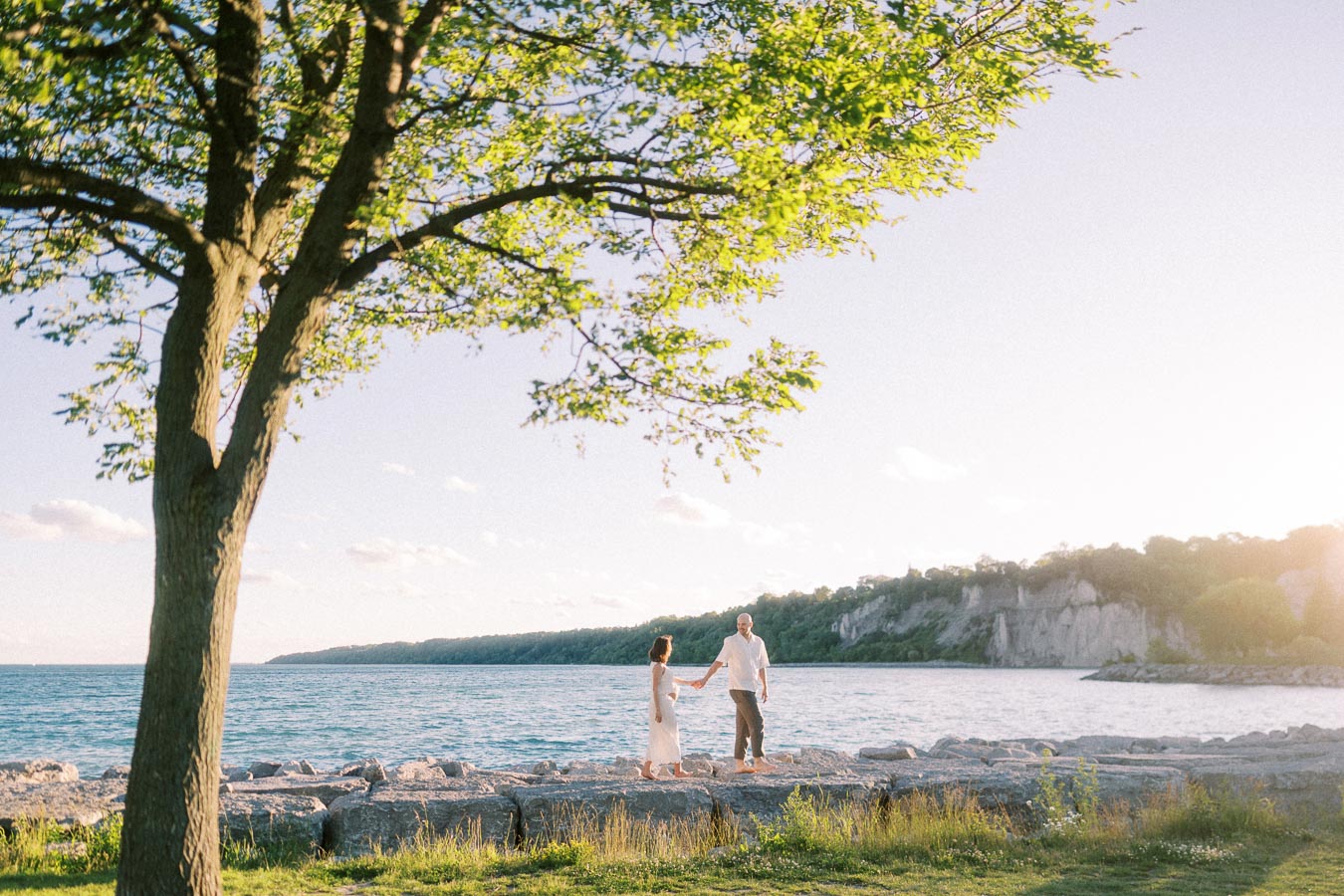Expecting couple holds hands on rocks beside Lake Ontario at Scarborough Bluffs.