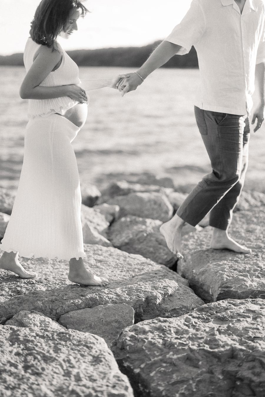 Pregnant woman in a white dress holds hands with her partner while standing barefoot on lakeside rocks.