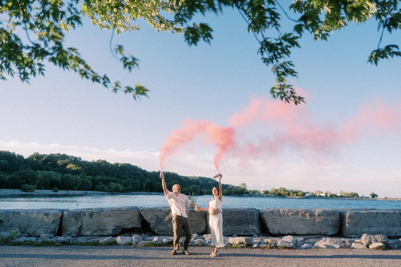 Expectant couple holding hands and raising pink smoke flares beside Lake Ontario rocks.