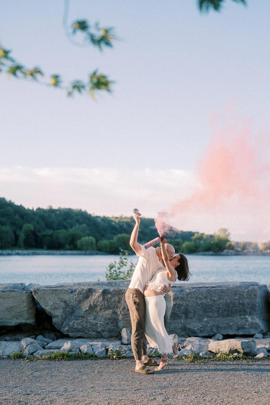 Expectant couple kisses while holding pink smoke by rocks and Lake Ontario.