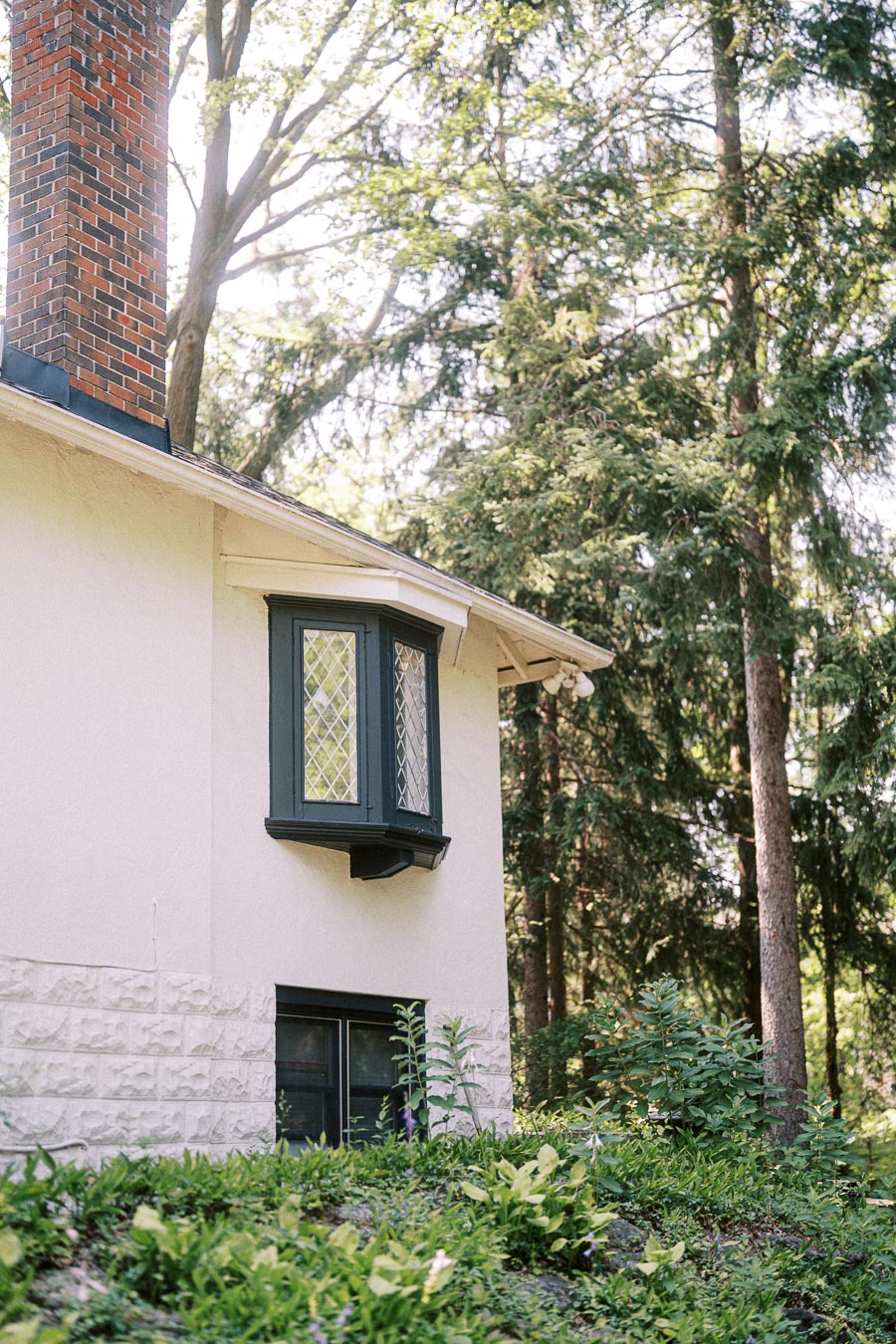 Cream house with brick chimney surrounded by trees and green garden plants.