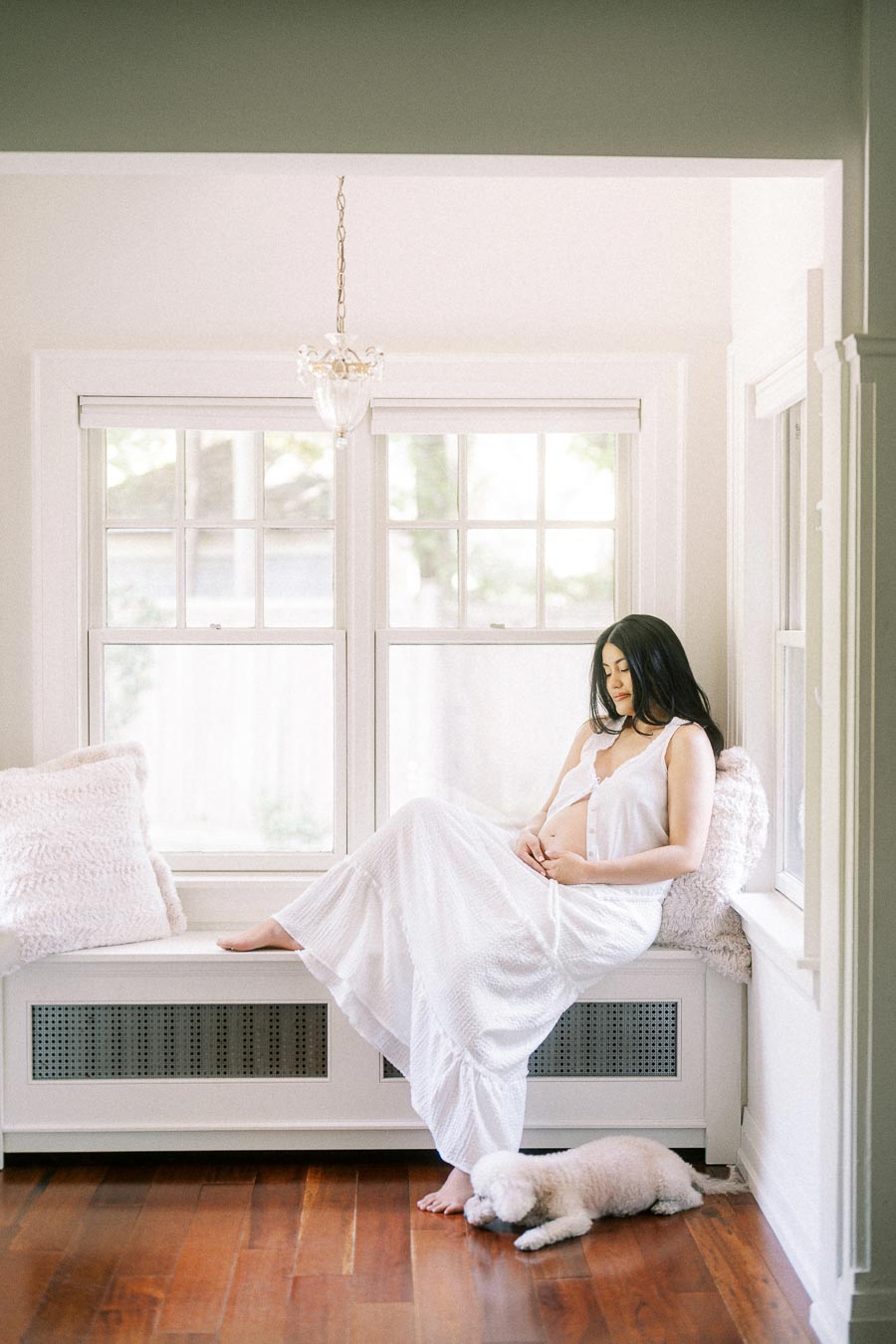 Pregnant woman in a white dress sitting by a bright window with a small dog nearby.