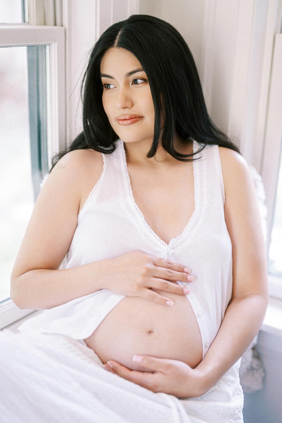 Pregnant woman in white clothing cradling her belly beside a bright window.