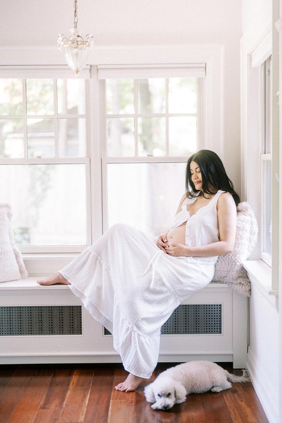 Pregnant woman in a white dress sitting by bright windows with a small dog nearby.