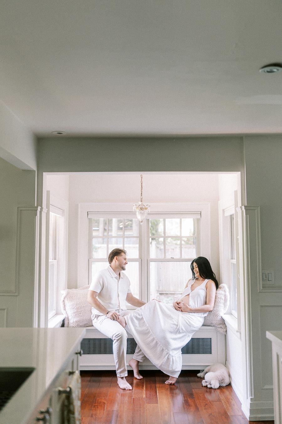 Couple in white outfits sitting together on a bright window bench indoors.