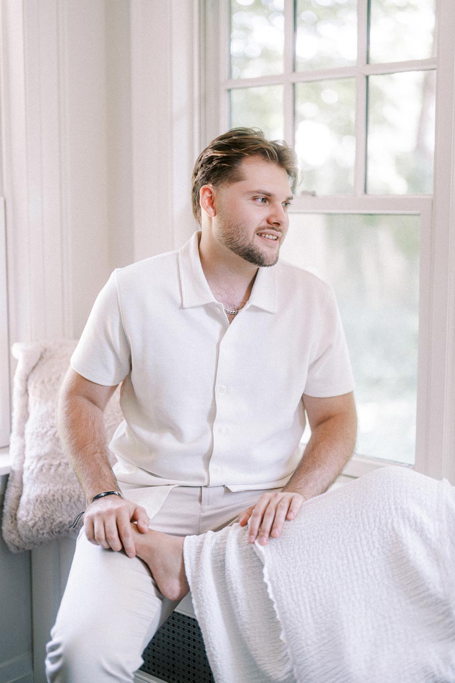 Man in white outfit seated by a bright window during an engagement session.