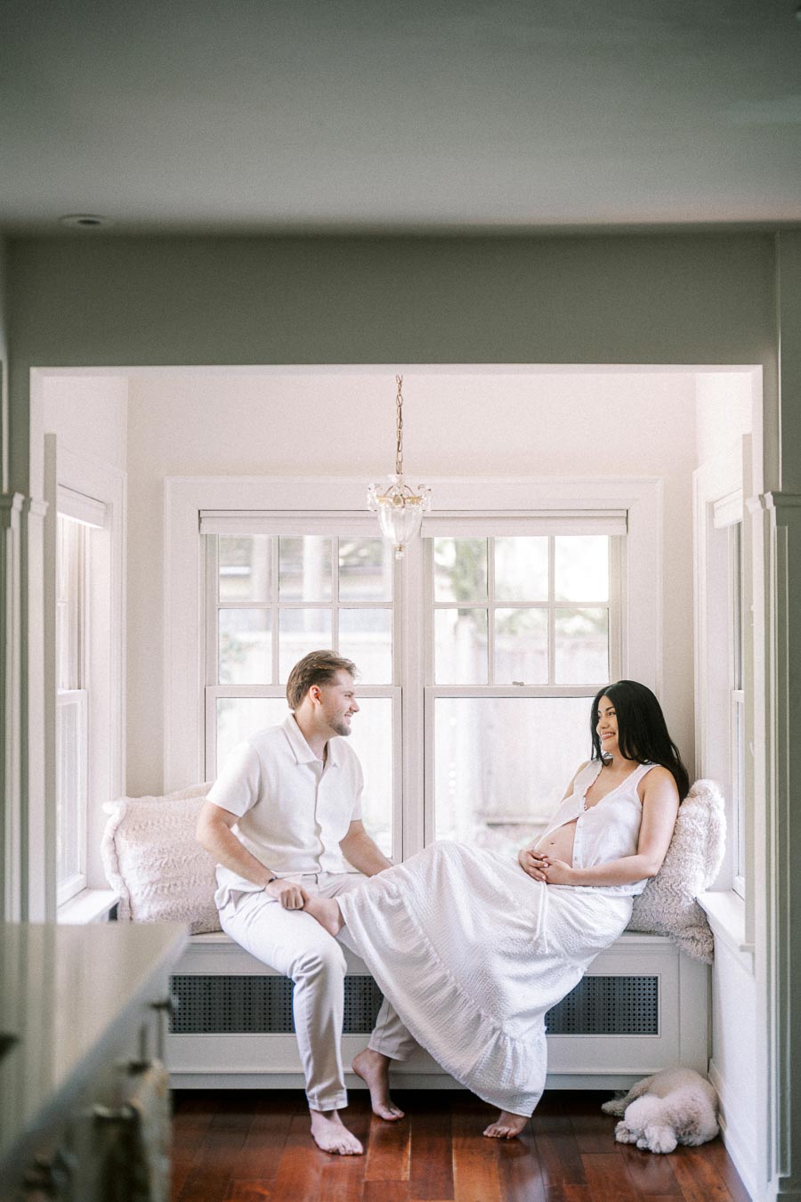 Couple in white outfits sitting together on a bright indoor window bench.
