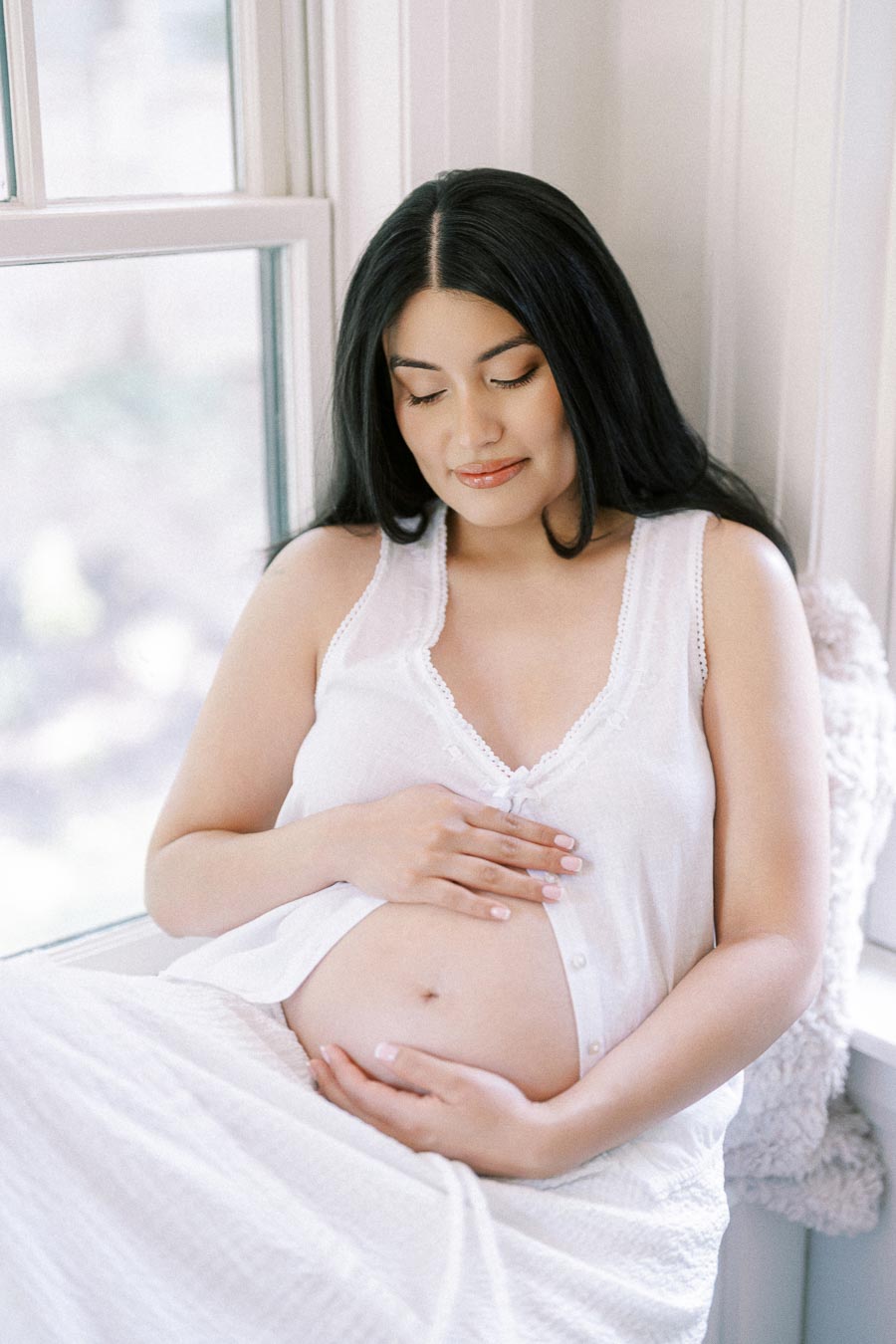 Pregnant woman in white sitting by a window with hands on her belly.
