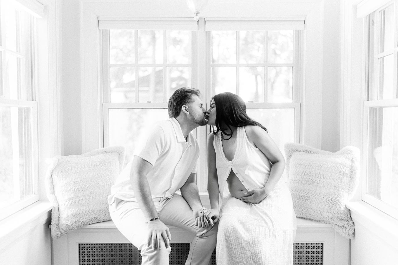 Couple in white outfits kissing while seated in a bright window nook.