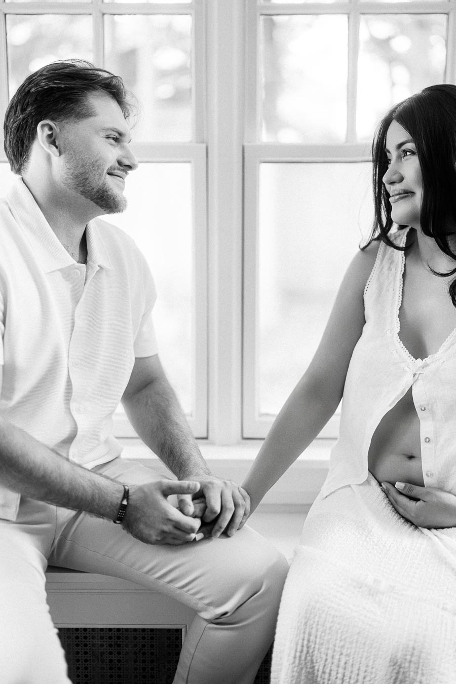 Couple holding hands and looking at each other beside a bright window.
