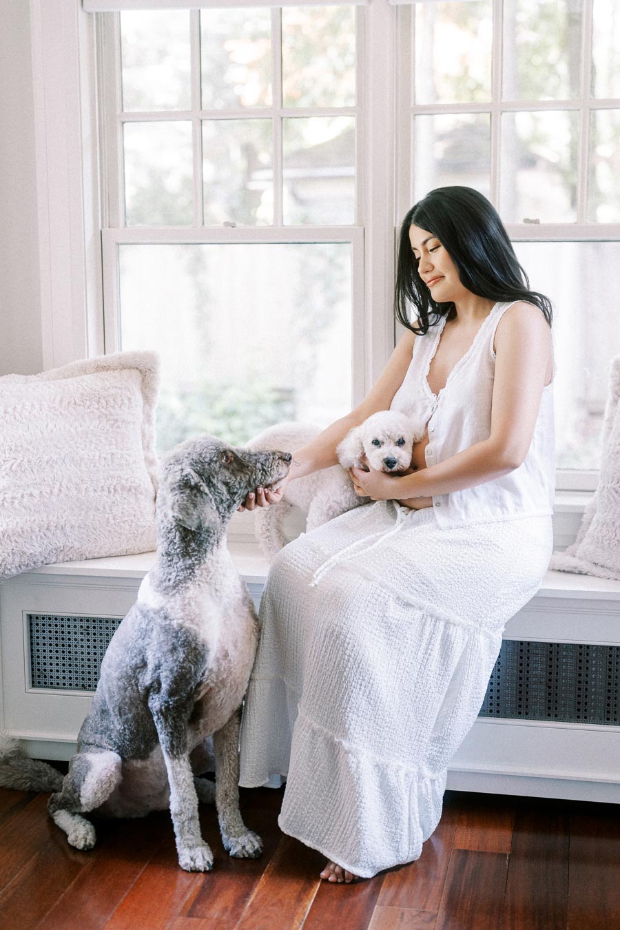 Woman in white dress sitting by windows with two small dogs.