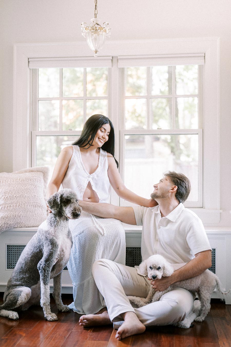 Couple in white outfits sitting indoors with two dogs by bright windows.