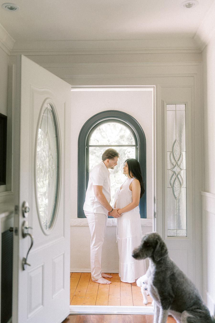 Couple in white holding hands by an arched window with a dog nearby.