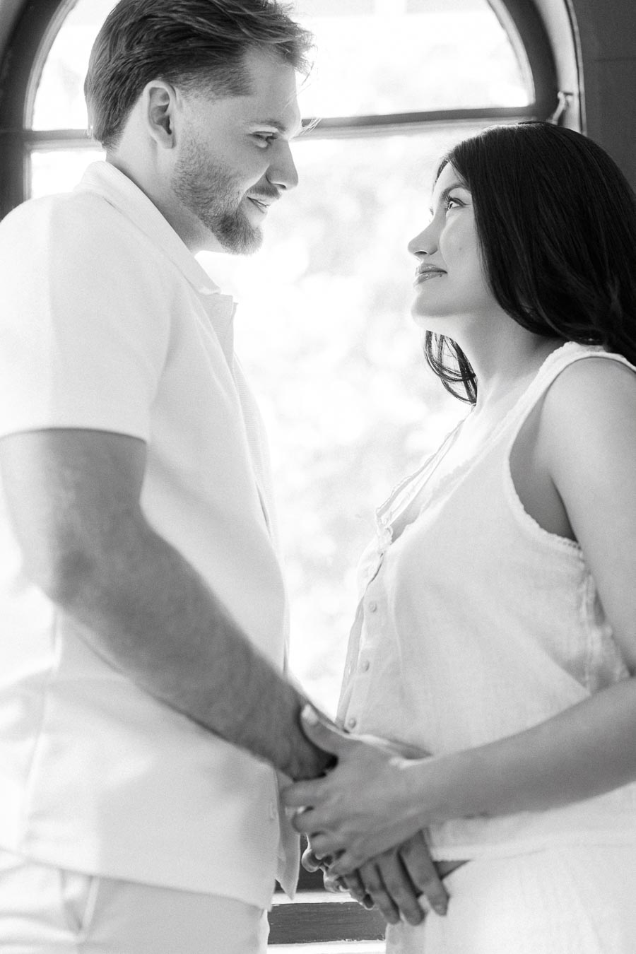 Couple in white clothing holding hands by an arched window.
