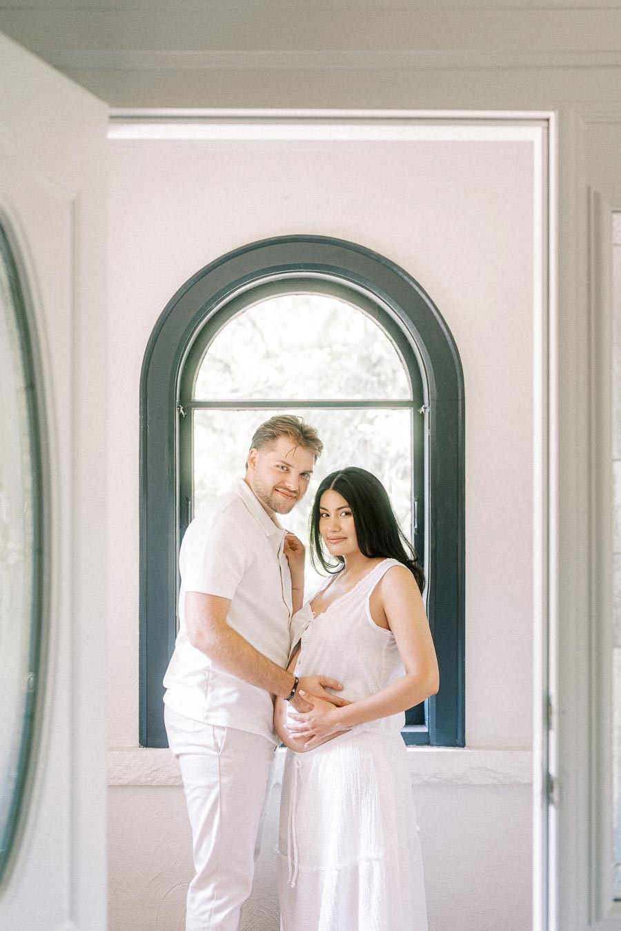 Couple in white outfits posing together beside an arched indoor window.
