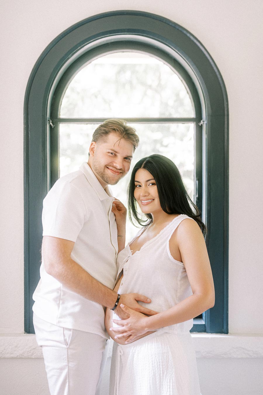 Couple in white outfits posing together by an arched window.