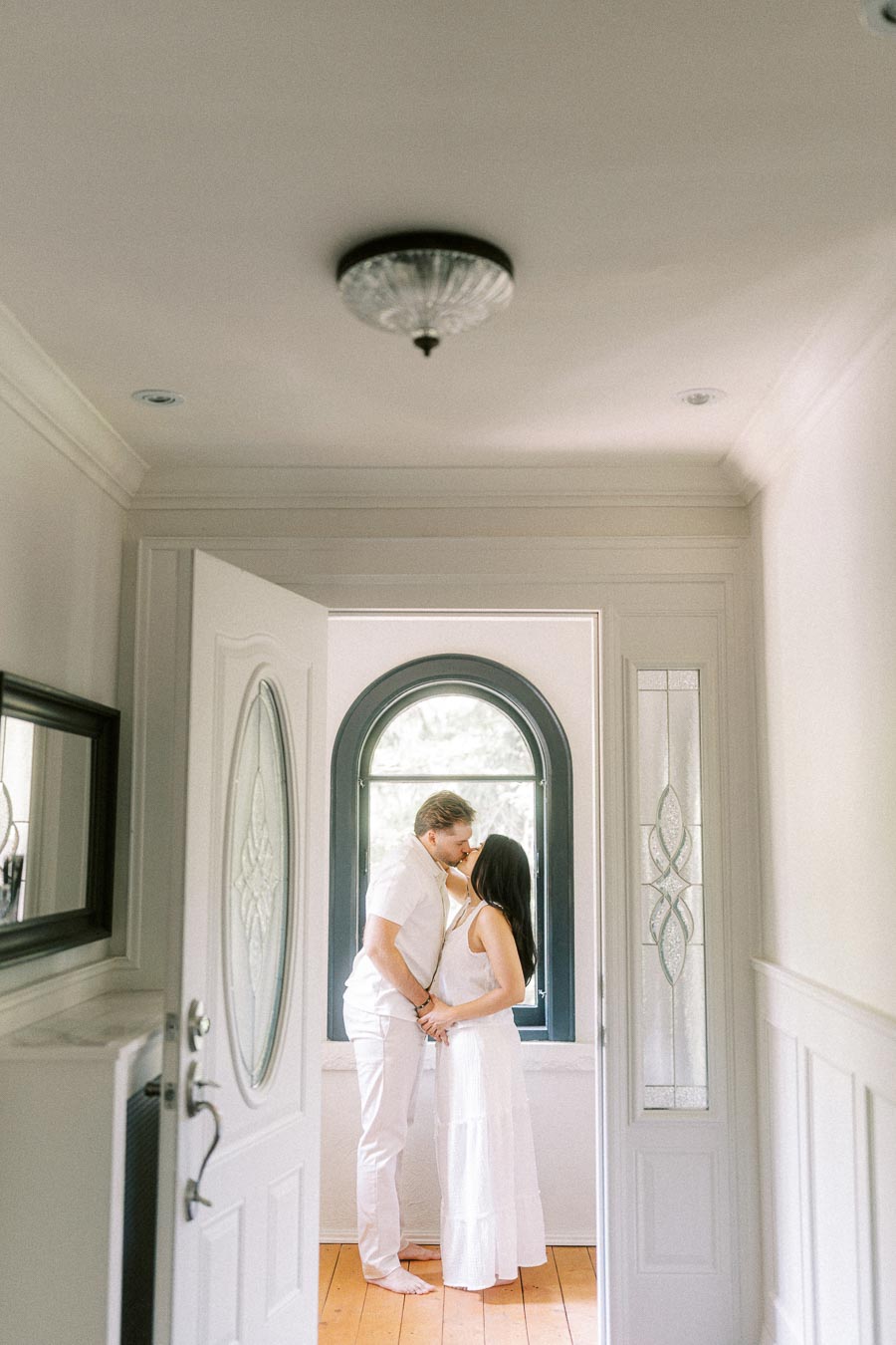 Couple in white outfits kissing by an arched window inside a bright entryway.