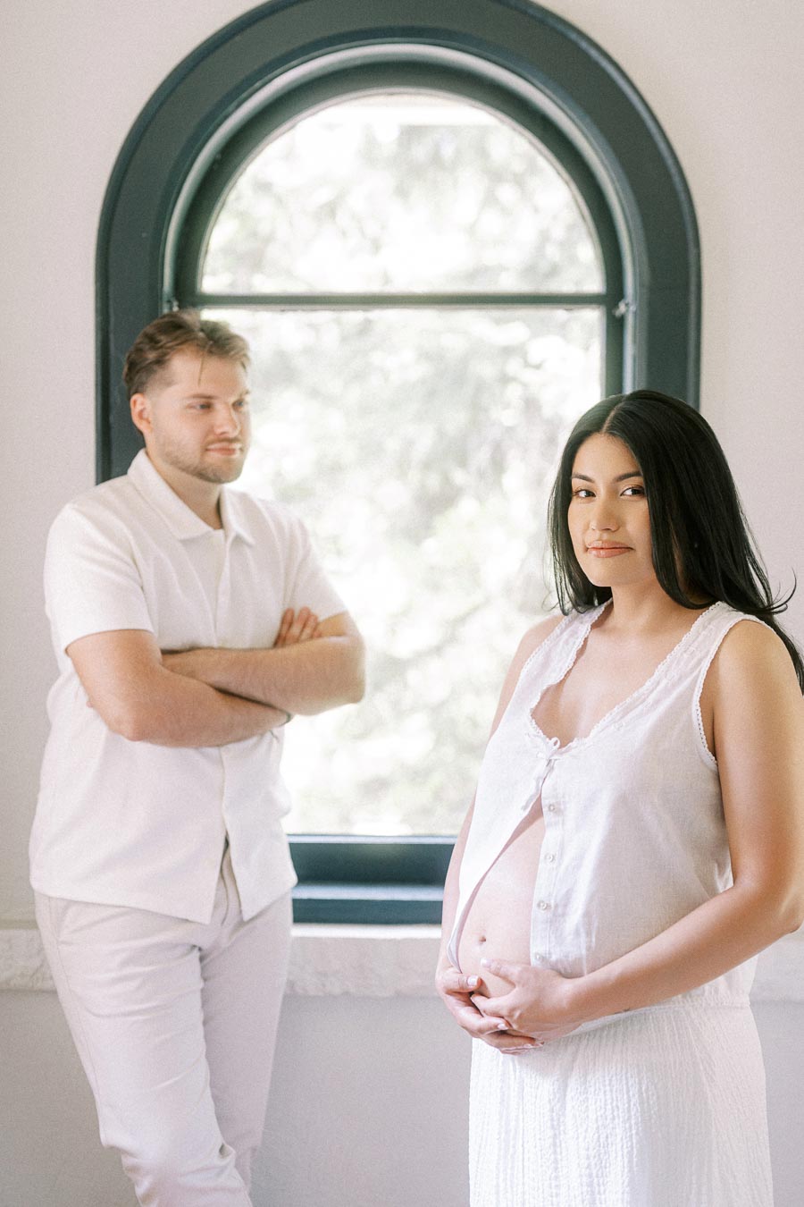 Expectant couple in white outfits posing by a tall arched window indoors.