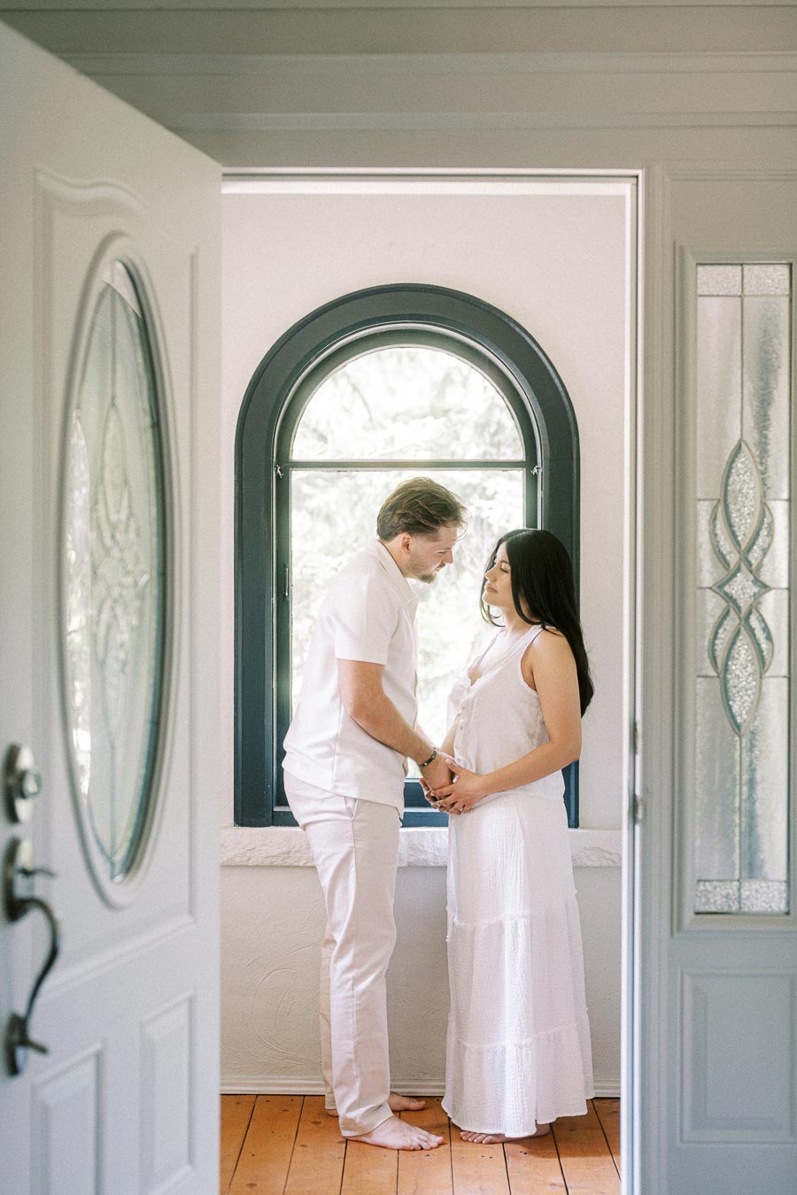 Couple in white outfits holding hands beside an arched indoor window.