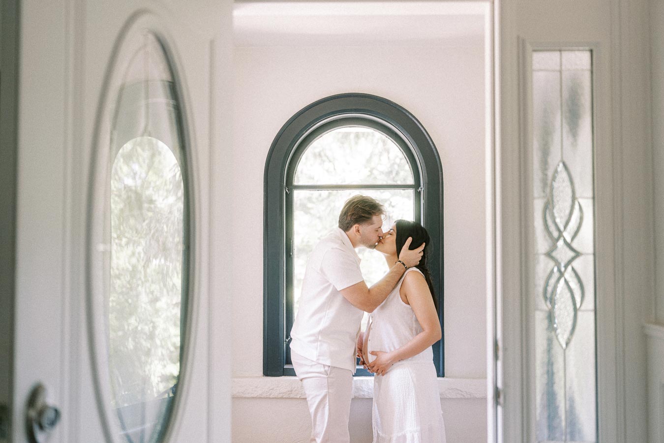 Couple in white outfits kissing beside an arched window indoors.