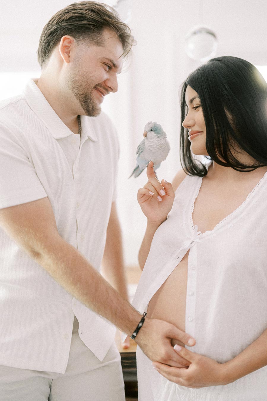 Expectant couple in white clothing smiling at a small bird indoors.