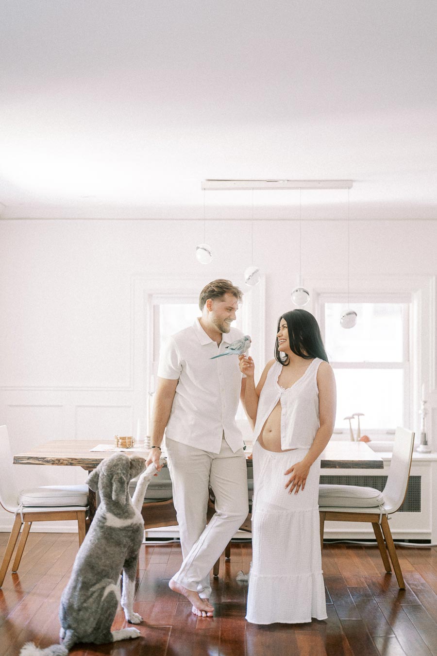 Expectant couple in white outfits standing indoors with their dog nearby.