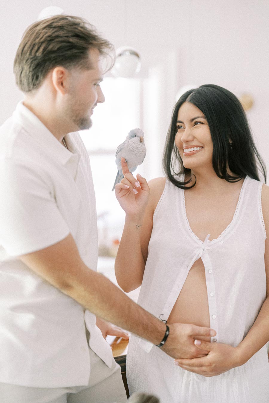 Expectant couple smiling indoors while a small grey parrot perches on her finger.