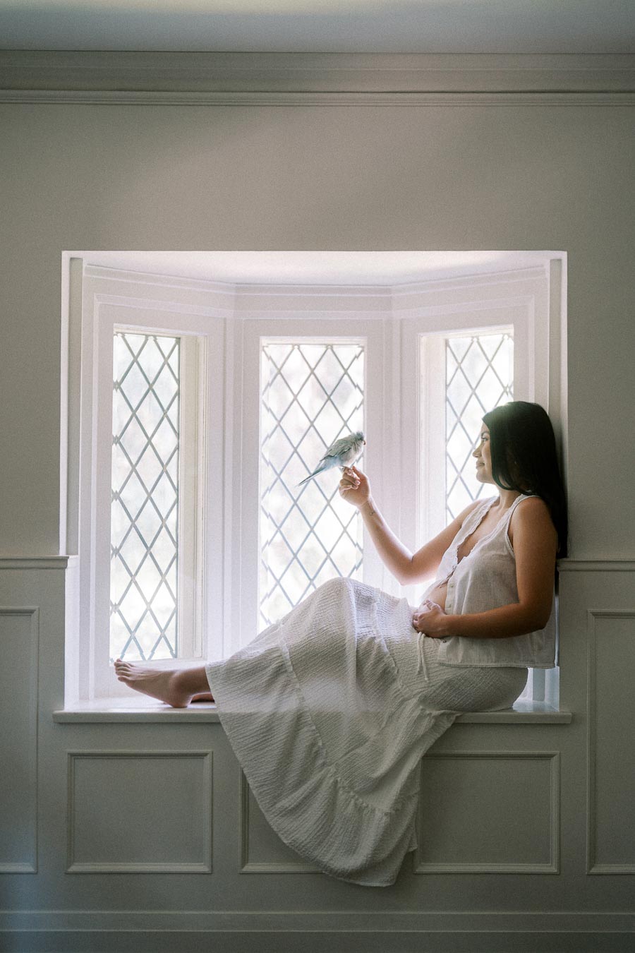 Woman in a white dress sitting in a window seat holding a small bird.