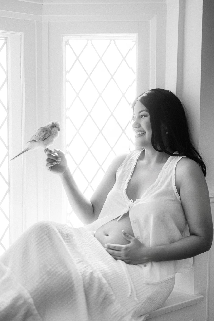 Pregnant woman in white sitting by a bright window holding a small bird.