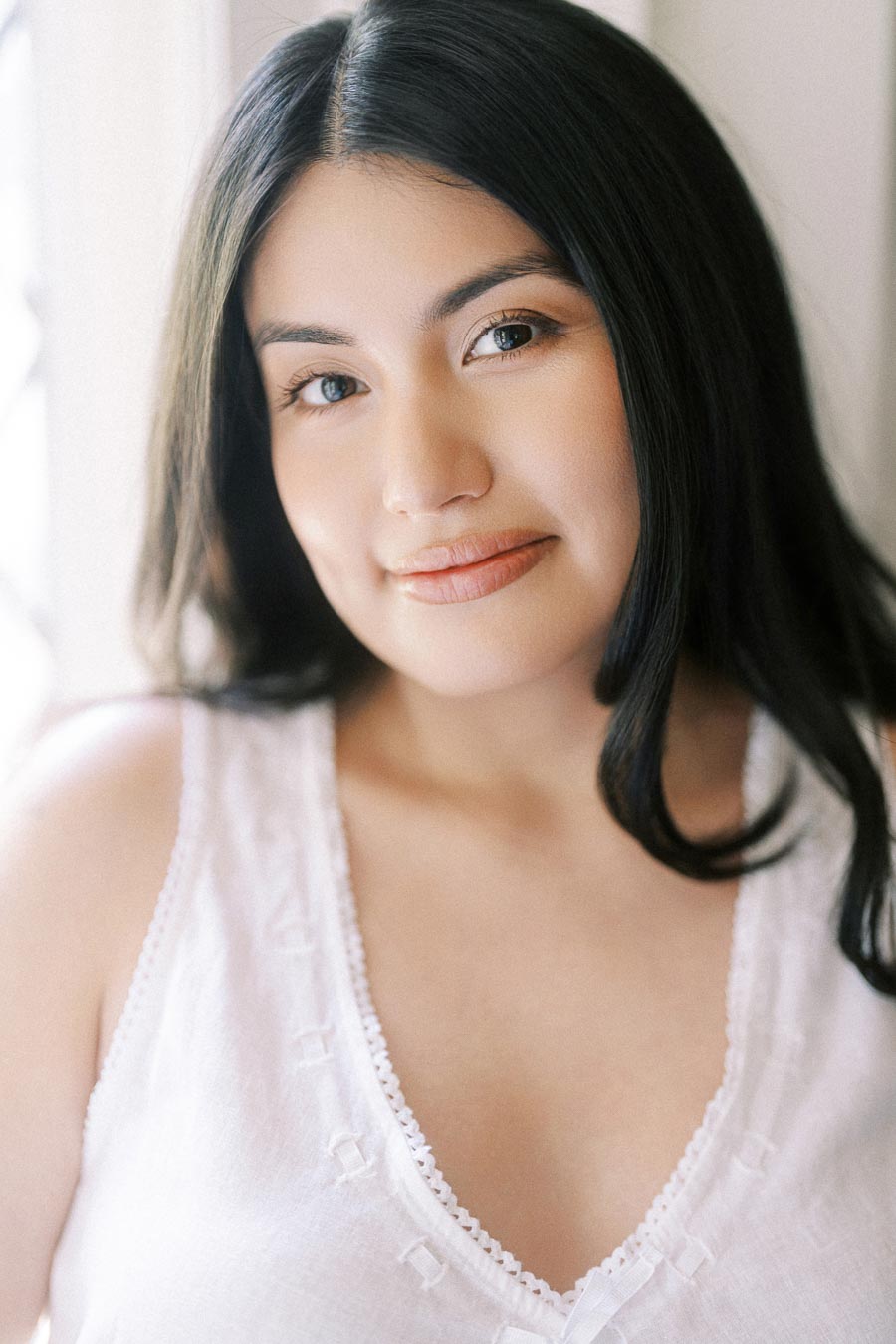 Close-up engagement portrait of a woman in a white top by soft window light.