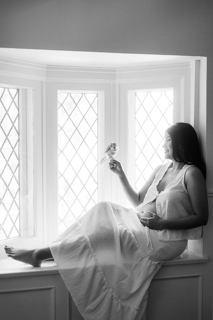 Pregnant woman sitting in a bright window seat holding a small flower.