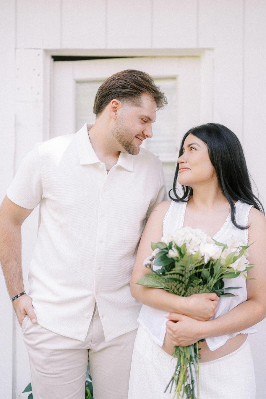 Couple in white outfits looking at each other as she holds a bouquet.