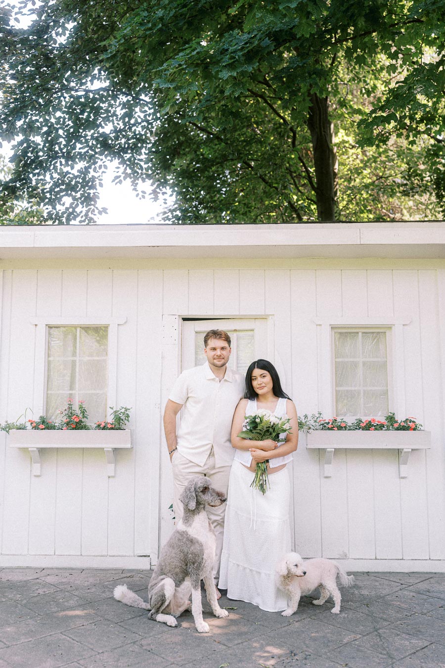 Engaged couple in white outfits posing with two dogs outside a white garden building.