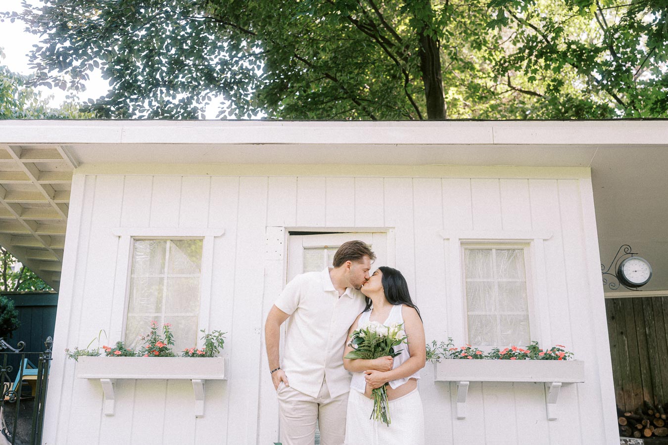 Engaged couple kissing outside a white garden building, holding a bouquet.