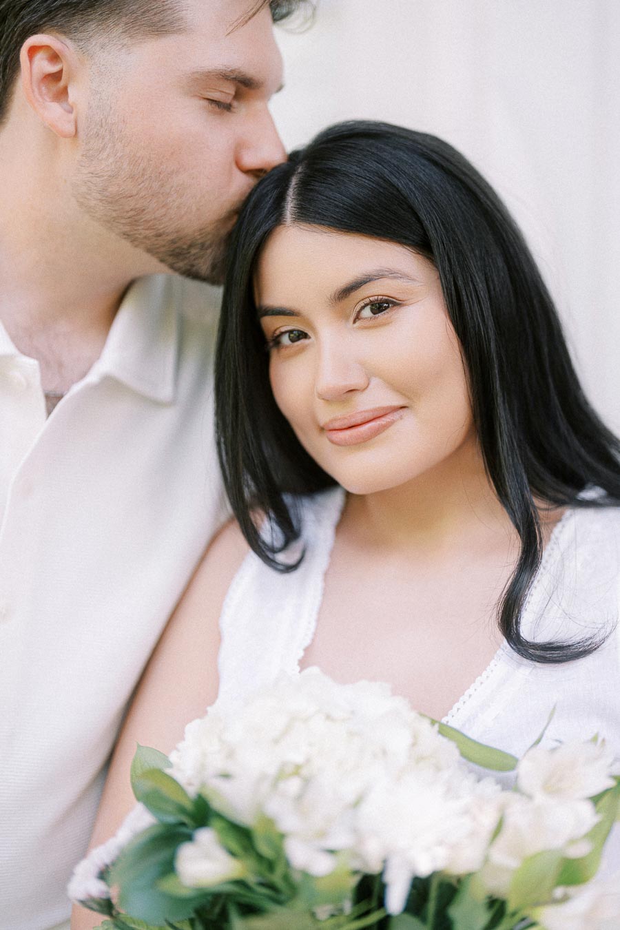 Close-up engagement portrait of a man kissing a woman’s forehead as she holds white flowers.
