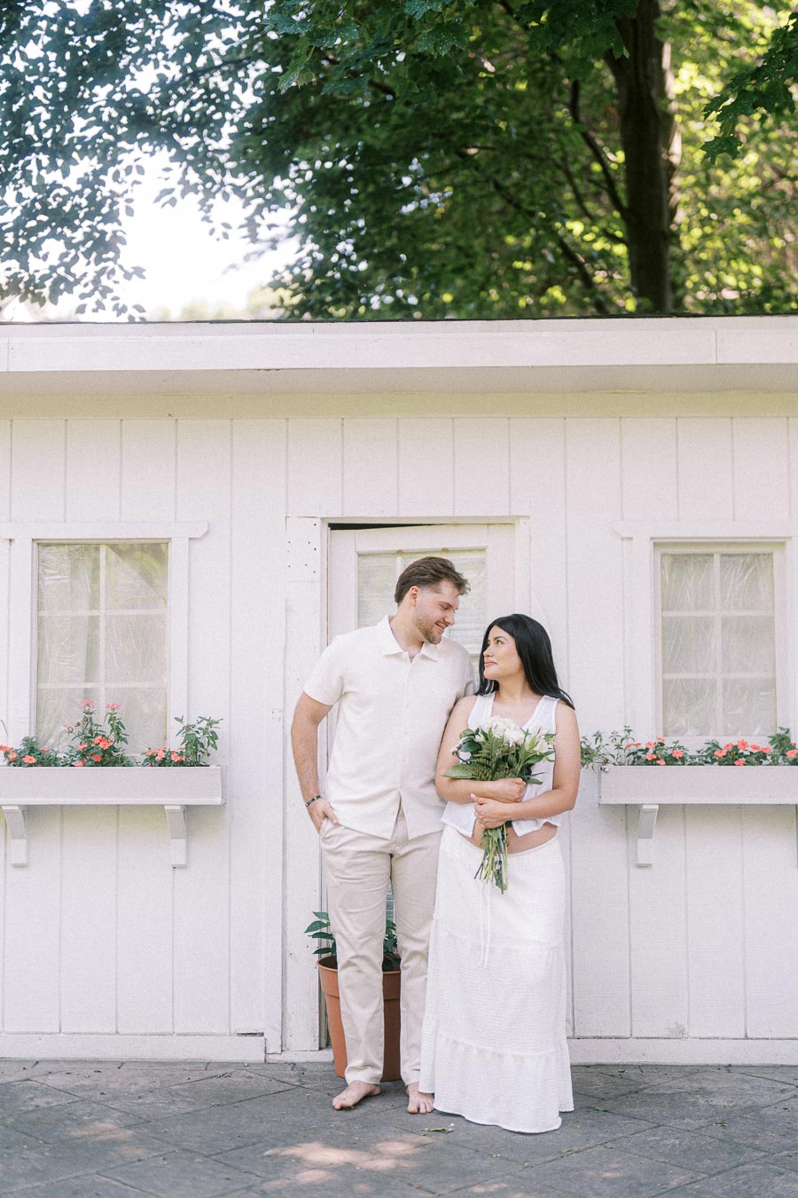 Engaged couple in white outfits standing by a white cottage with flowers.