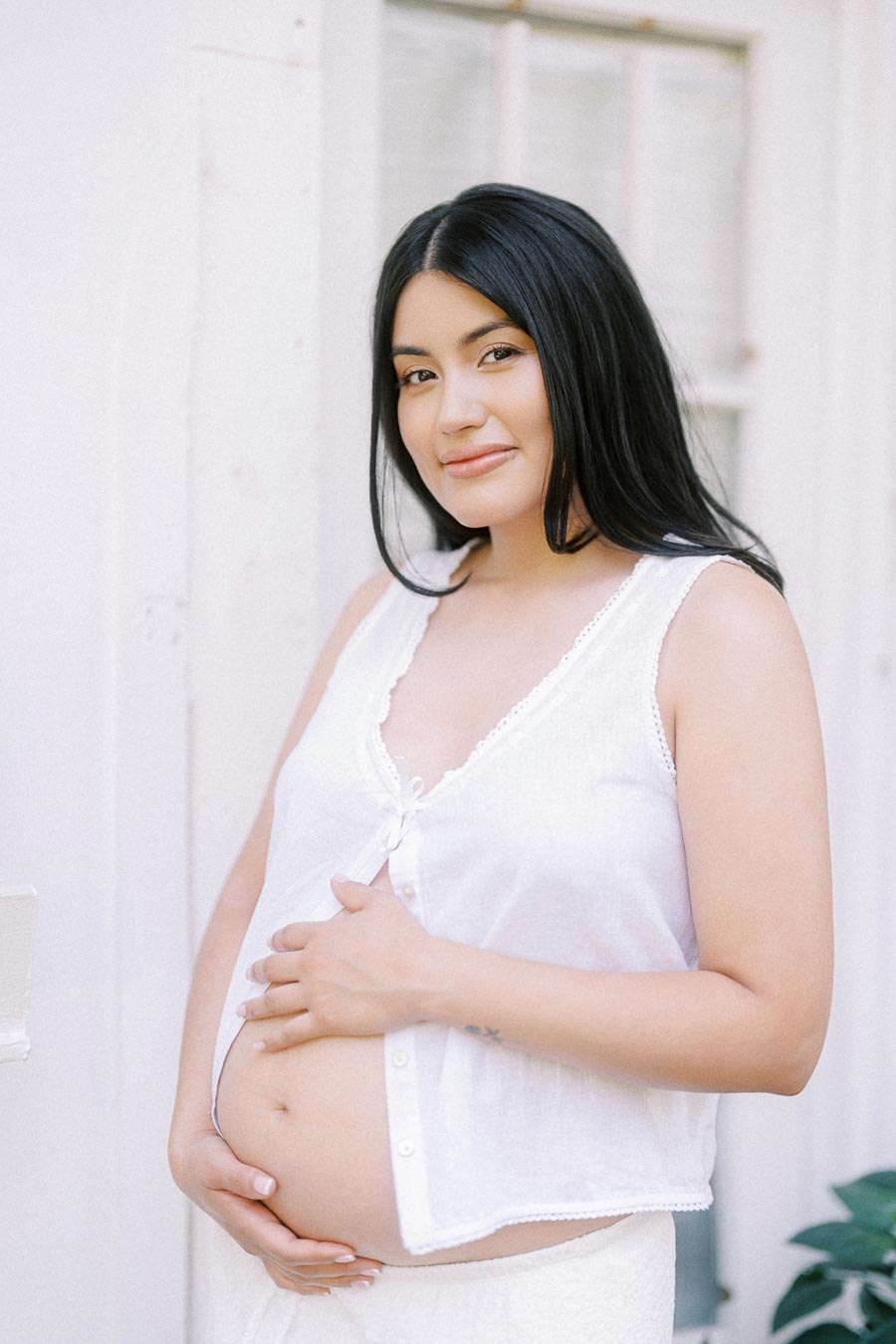 Pregnant woman in white clothing gently holding her belly by a bright window.