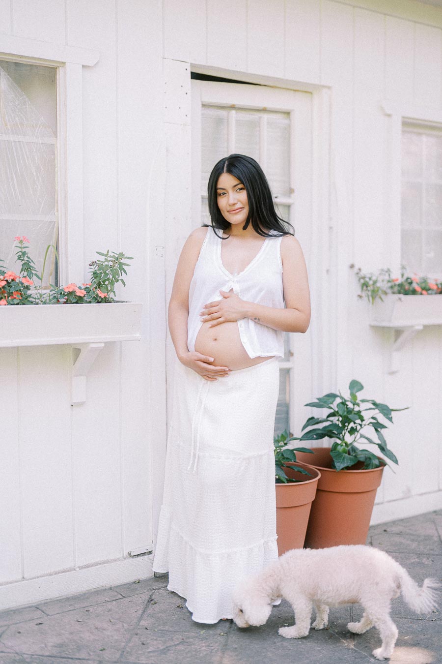 Pregnant woman in a white outfit standing by potted plants with a small dog.