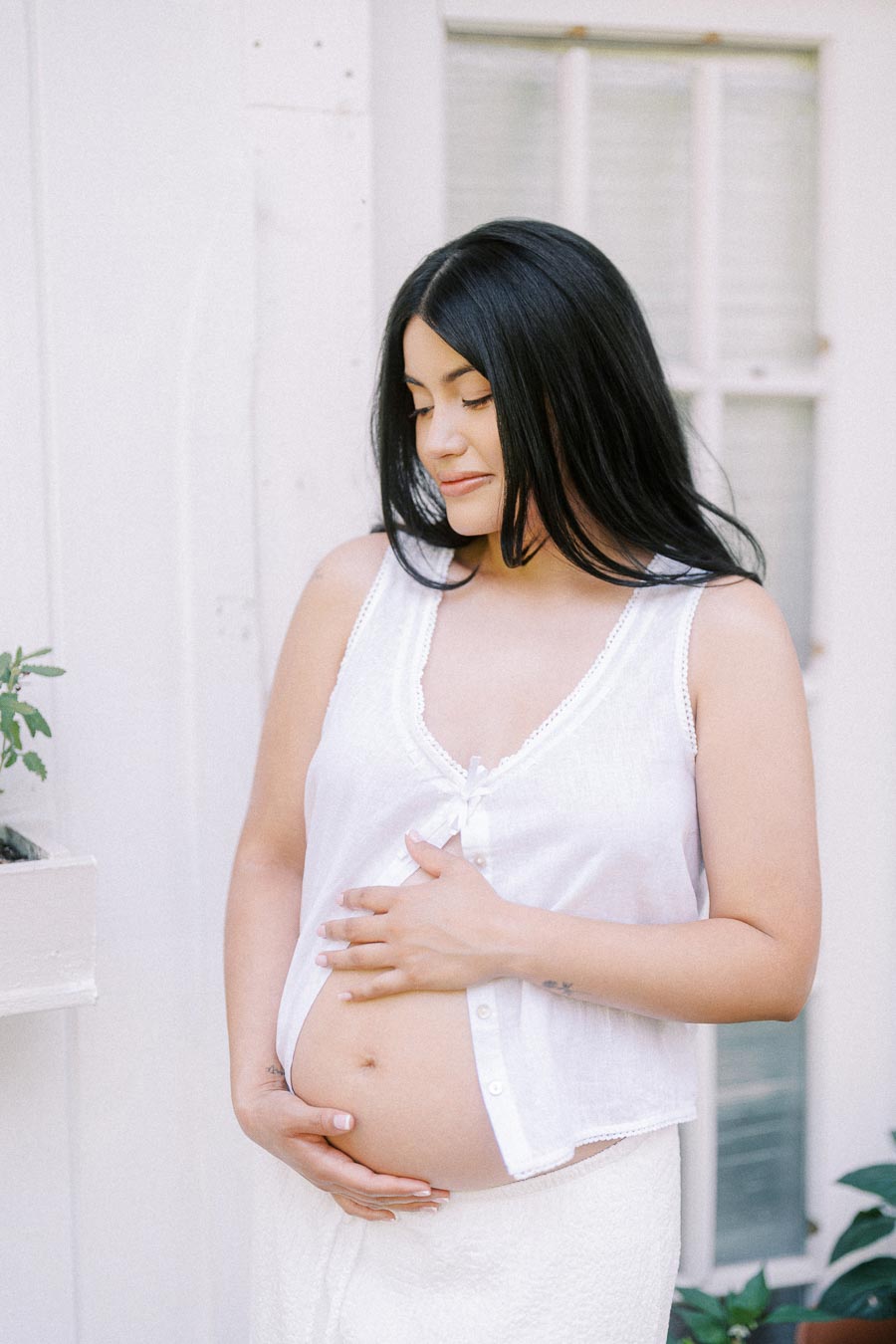 Pregnant woman in white clothing cradling her belly beside a window.