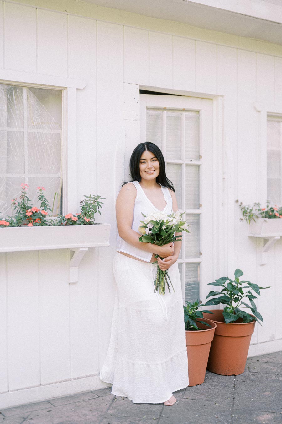 Woman in white outfit holding flowers beside a white building with potted plants.
