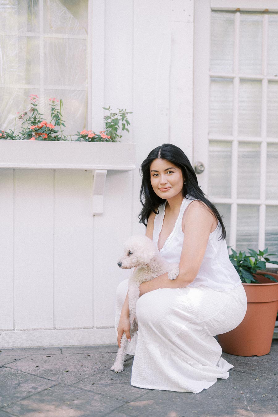 Woman in a white dress crouching beside a small white dog on a patio.
