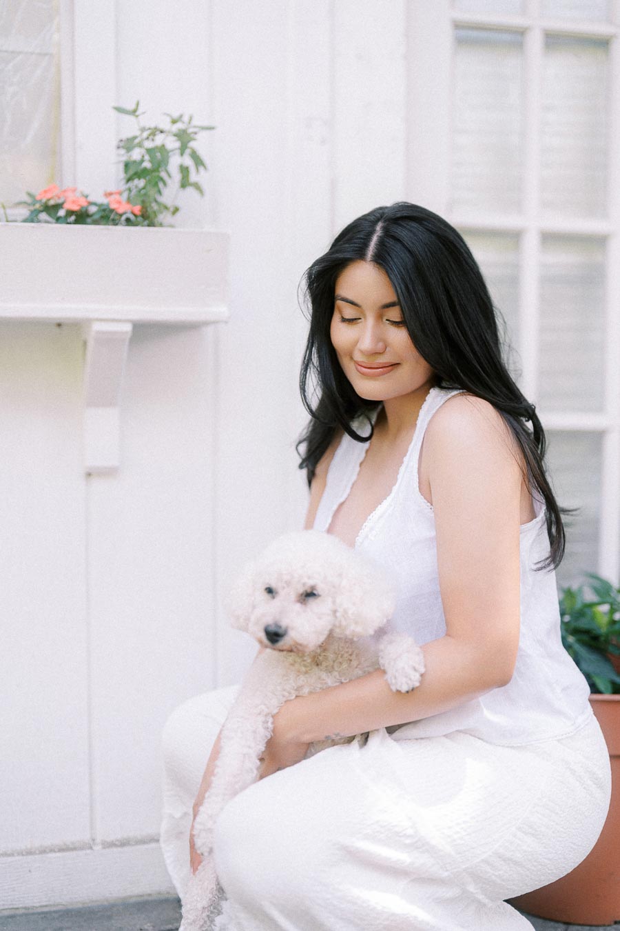 Woman in white holding a small white dog beside a pale building.