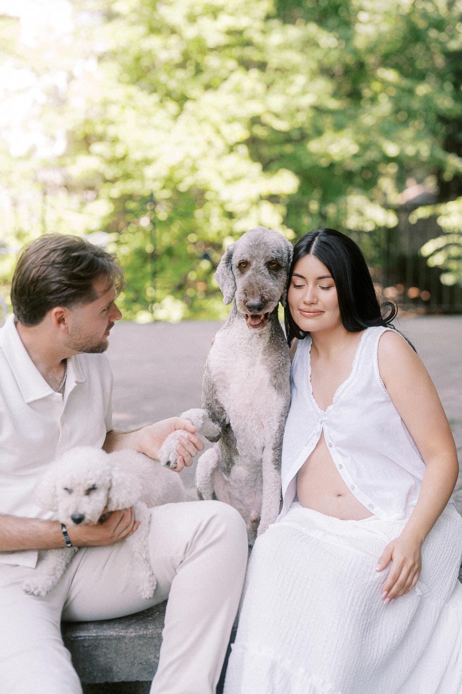 Couple in white outfits sitting outdoors with two dogs during an engagement photo.