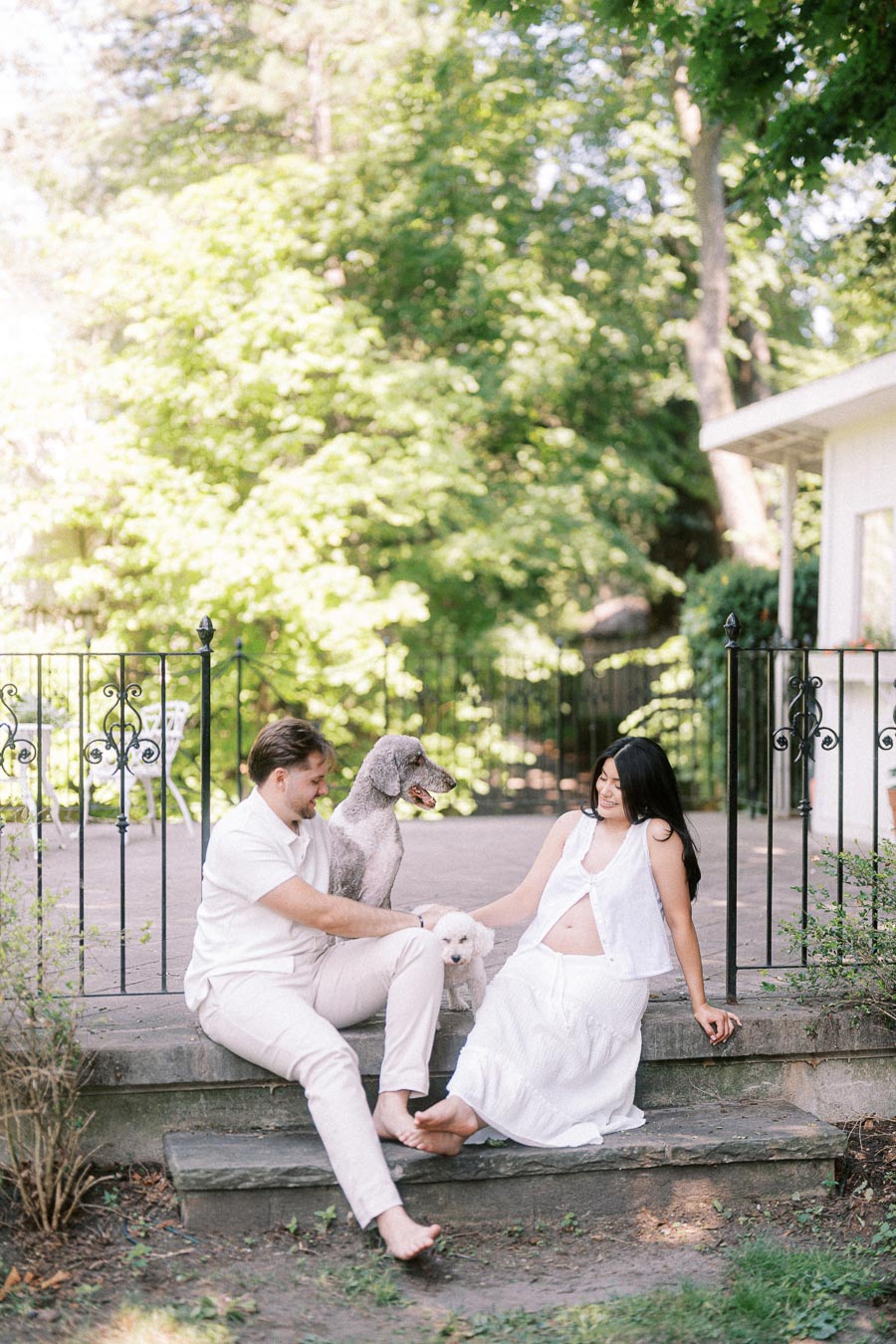 Couple in white sitting on garden steps with two dogs during engagement photo.