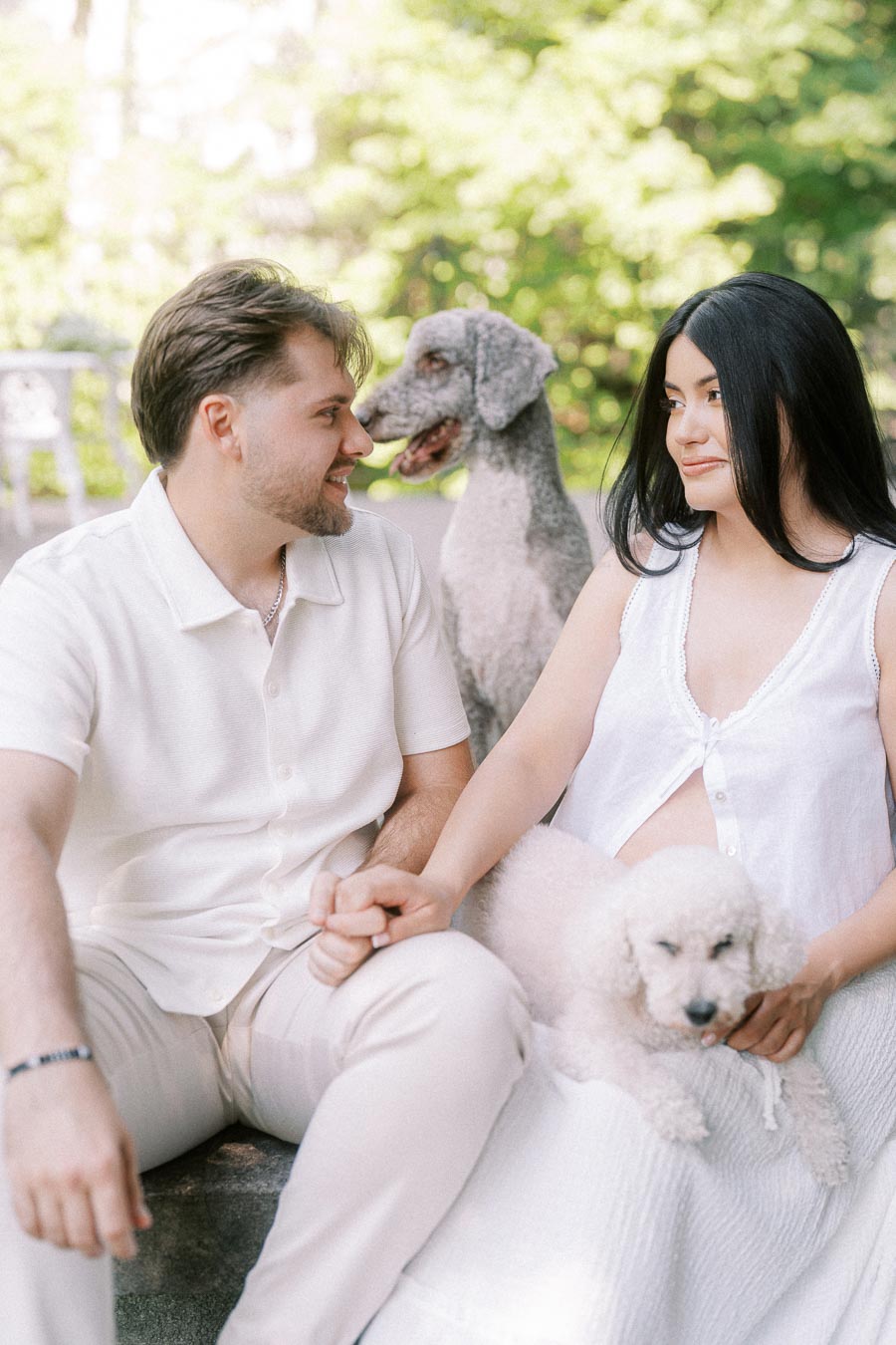 Engaged couple seated outdoors holding hands with two poodles beside them.