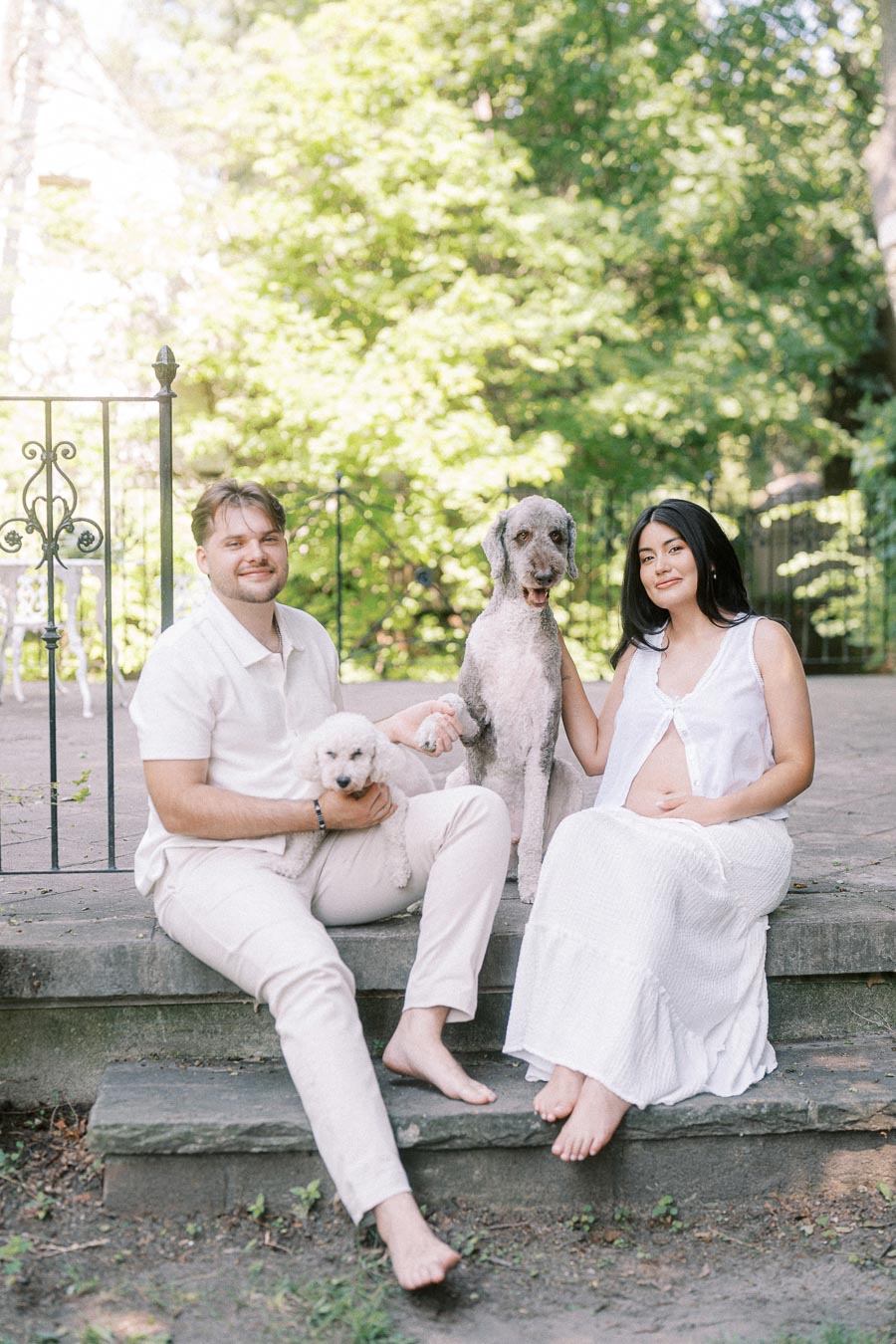 Engaged couple in white sitting on stone steps with two dogs in a garden.