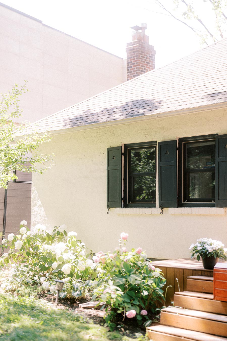 Sunlit house exterior with green shutters, garden flowers, and wooden steps.